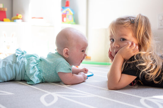 Two sisters laying on a floor during tummy time. Toddler and new baby. Shocked toddler. New baby in family.