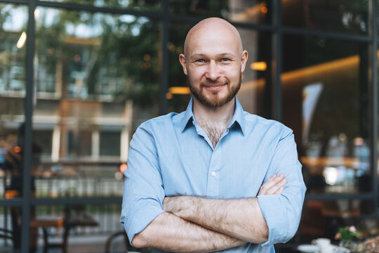 Portrait of Adult bald smiling attractive man forty years with beard in blue shirt businessman against glass wall of the street cafe