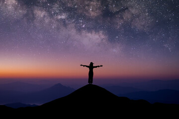 Silhouette of young female traveler wearing hat standing and open arms watched the beautiful night sky, star and milky way alone on top of the mountain.