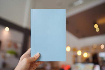 Woman hand holding a light blue book with blank cover for inserting text on blurred cafe background.