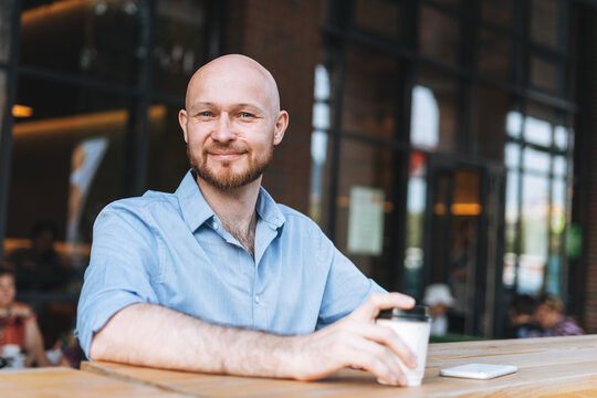 Adult bald smiling attractive man forty years with beard in blue shirt businessman using mobile phone with paper cup of coffee at cafe