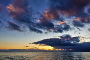 Colorful sunset over the sea. There are purple and pink clouds in the blue sky. Orange glow over the horizon. The water is calm. Reflection. 