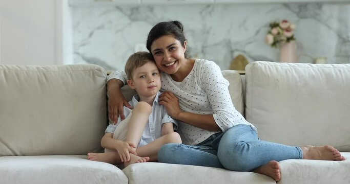 Attractive Indian woman sit on sofa with little preschooler, cute Caucasian boy smiling looking at camera. New loving mother for adopted child, custody and foster, multi racial family portrait concept
