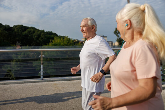 Smiling mature man and woman with ponytail run together along footbridge on summer day