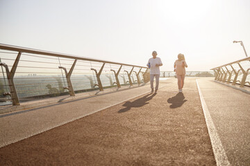 Positive senior woman and man with bottles run along footbridge in summer evening