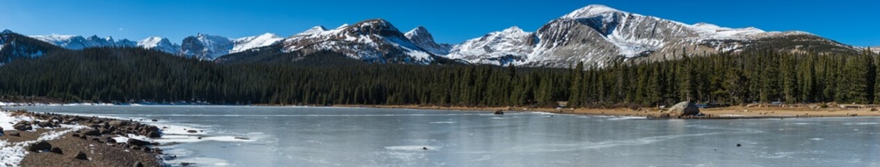 A wide angle panoramic landscape shot of snowy mountains, a line of pine trees, and a frozen lake and shore line panorama - Powered by Adobe