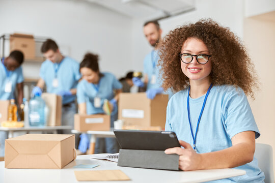 Attractive young woman, volunteer in blue uniform using tablet pc and smiling at camera while sitting indoors. Team sorting, packing items in the background