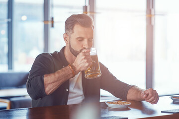 Handsome young man in casual clothing drinking beer while spending time in the pub