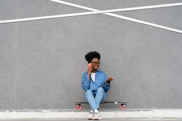 Joyful trendy black girl talking on phone call. Stylish african woman in urban denim outfit calling and communicating using cell phone outdoors. Mobile connection, technology and lifestyle concept