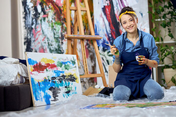 Obraz na płótnie Canvas Joyful female painter in apron smiling at camera, holding paintbrush while working on painting, sitting on the floor at home studio workshop