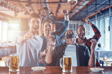 Excited young men in casual clothing drinking beer and watching sport game while sitting in the pub