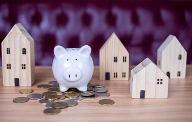 A white piggy bank is placed on a pile of coins. And next to it is a wooden model house. On the background is a red sofa. all on a wooden table