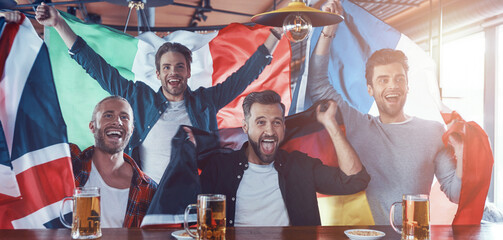 Excited young men covered in international flags drinking beer and watching sport game while sitting in the pub