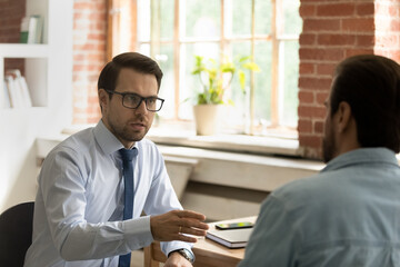 Obraz premium Serious male colleagues discuss business seated at desk. Two business partners negotiating meet in office, search together problem solution, share decision, express opinion, communicate at workplace