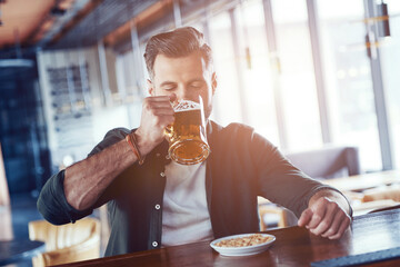 Charming young man in casual clothing drinking beer while spending time in the pub