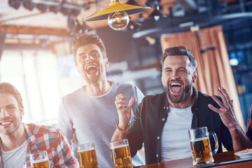 Cheering young men in casual clothing drinking beer and watching sport game while sitting in the pub