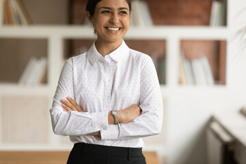 Portrait of smiling Indian businesswoman stands in modern office with arms crossed look into...