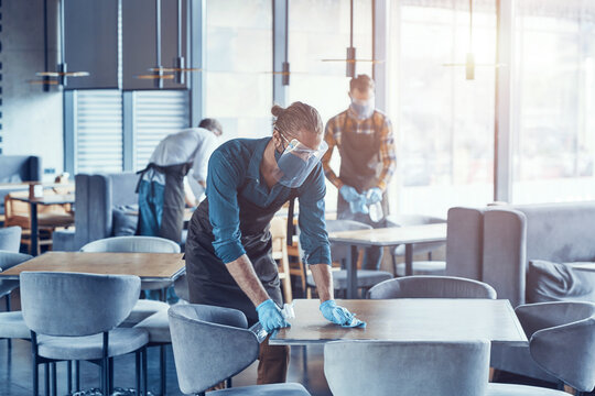 Three Young Male Waiters In Protective Workwear Cleaning Tables For Clients In Restaurant