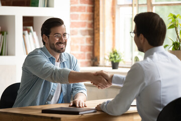 Two confident business partners sit at desk shake hands after successful formal meeting. Applicant position candidature greets HR manager start job interview, make deal, negotiation, agreement concept