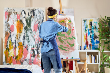 Obraz premium Back view of young female artist in apron creating abstract painting on easel, applying paint on canvas with fingers, standing in living room at home studio