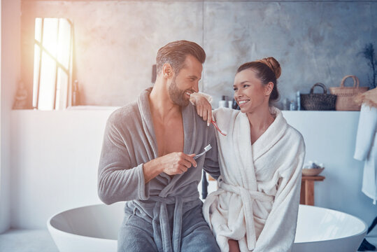 Beautiful young couple in bathrobes smiling and cleaning teeth while doing morning routine