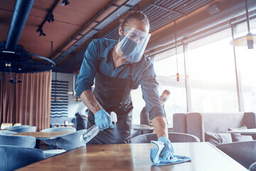Young male waiters in protective workwear cleaning tables in restaurant