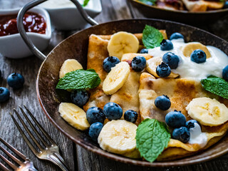 Sweet crepes with bananas, strawberries jam and blueberries in pan on wooden table
