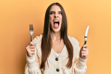 Beautiful brunette young woman holding fork and knife ready to eat angry and mad screaming frustrated and furious, shouting with anger looking up. - Powered by Adobe