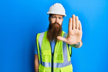 Redhead man with long beard wearing safety helmet and reflective jacket doing stop sing with palm of the hand. warning expression with negative and serious gesture on the face. - obrazy, fototapety, plakaty