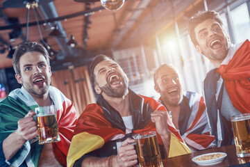 Happy young men covered in international flags drinking beer and watching sport game while sitting...