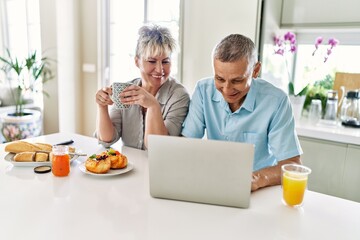 Senior caucasian couple having breakfast using laptop at the kitchen.