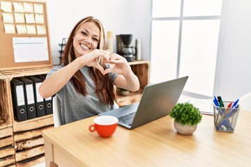 Young brunette woman working at the office with laptop smiling in love doing heart symbol shape with hands. romantic concept.