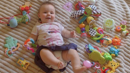 The little girl lies on the bed wearing toys. There are items for independent play nearby. She moves her legs. Relaxation of the child's mood.