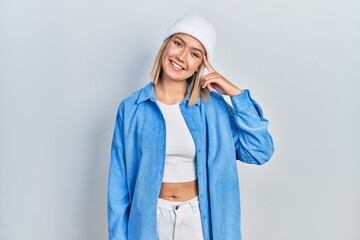 Beautiful blonde woman wearing wool hat smiling pointing to head with one finger, great idea or...