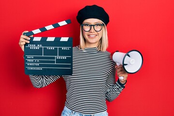 Beautiful blonde woman holding video film clapboard and megaphone smiling with a happy and cool...