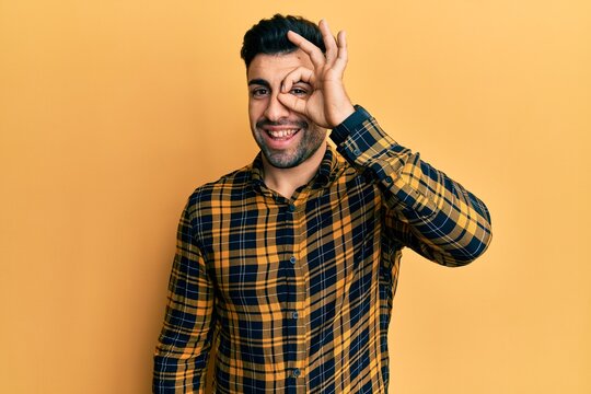 Young hispanic man wearing casual clothes doing ok gesture with hand smiling, eye looking through fingers with happy face.