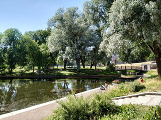 Fototapeta premium On a summer day, people relax in the garden. View of the trees, pond, bridge in the Yusupovsky Garden in the old center of St. Petersburg. Recreation places for residents of the city of St. Petersburg