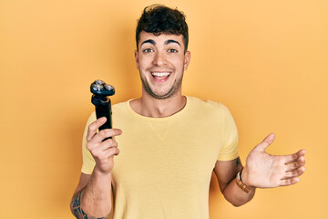 Young hispanic man holding electric razor celebrating achievement with happy smile and winner expression with raised hand - obrazy, fototapety, plakaty
