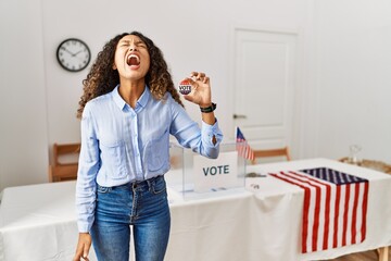 Beautiful hispanic woman standing by at political campaign by voting ballot angry and mad screaming frustrated and furious, shouting with anger. rage and aggressive concept.