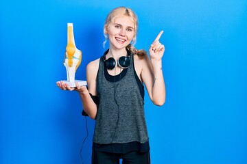 Beautiful caucasian sports woman with blond hair holding anatomical model of knee joint surprised with an idea or question pointing finger with happy face, number one