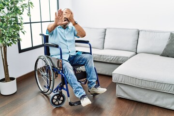 Handsome senior man sitting on wheelchair at the living room covering eyes with hands and doing...