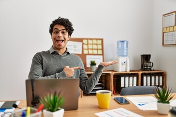 Fototapeta premium Young hispanic man wearing business style sitting on desk at office amazed and smiling to the camera while presenting with hand and pointing with finger.
