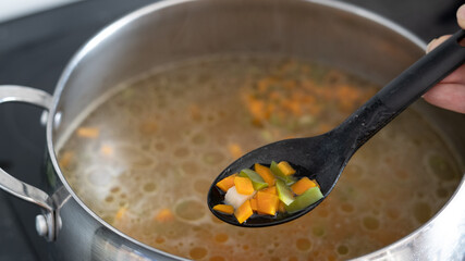 Vegetable soup in a saucepan: carrots, potatoes, peppers, beans, onions. vegetables preparing on an electric stove. The girl's hand holds a spoon with bright boiled vegetables.