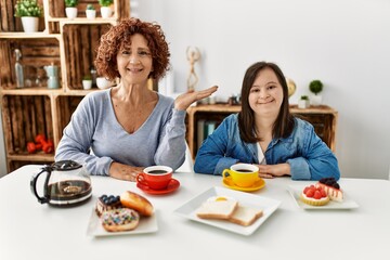 Family of mother and down syndrome daughter sitting at home eating breakfast smiling cheerful presenting and pointing with palm of hand looking at the camera.