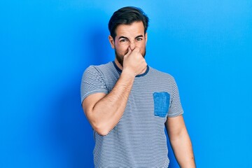 Handsome caucasian man with beard wearing casual striped t shirt smelling something stinky and...