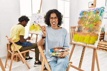 Young hispanic woman at art studio pointing aside worried and nervous with forefinger, concerned...