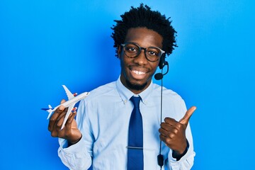 Young african american travel agent man holding plane smiling happy and positive, thumb up doing...