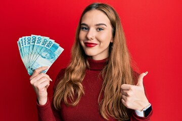 Young blonde woman holding 100 brazilian real banknotes smiling happy and positive, thumb up doing...