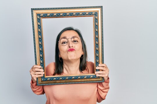 Middle age hispanic woman holding empty frame looking at the camera blowing a kiss being lovely and sexy. love expression.