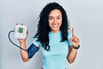 Young hispanic woman with curly hair using blood pressure monitor smiling with an idea or question...